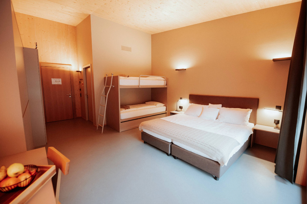 Camera Family del Campra Lodge: ambienti luminosi e spaziosi pensati per il comfort di famiglie e gruppi, con letto matrimoniale, arredi essenziali, balcone privato e vista rilassante sulla natura incontaminata dell’Alta Valle di Blenio